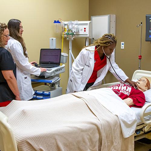 Three healthcare professionals practice medical procedures on a patient simulator in a hospital room. One uses a stethoscope, another operates a computer, and the third oversees the activities.