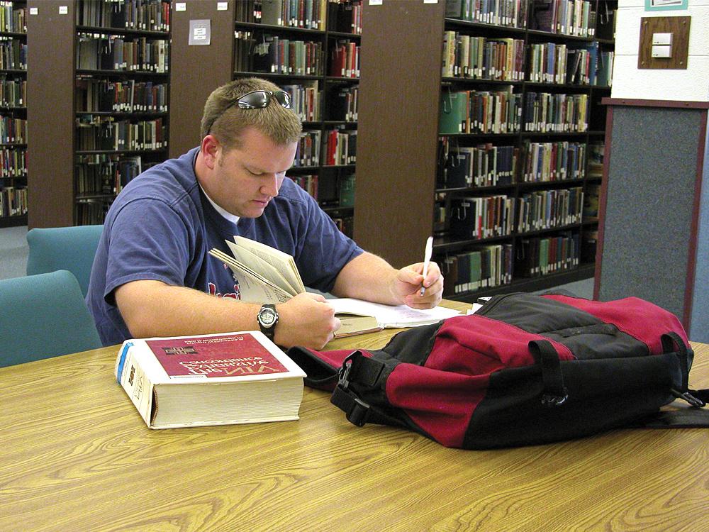 A person wearing a blue shirt is studying at a library table with a red and black backpack and an open book in front of them. 书架在背景中可见.